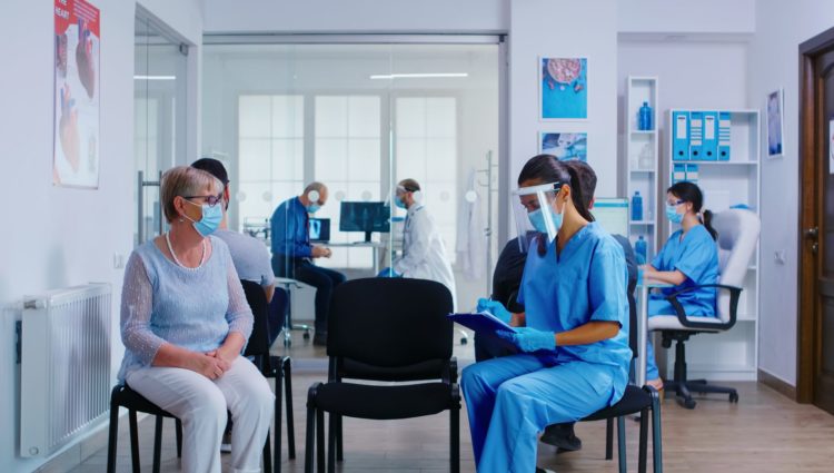 Hospital waiting room. Patient wearing a mask sits across from a nurse wearing a visor.