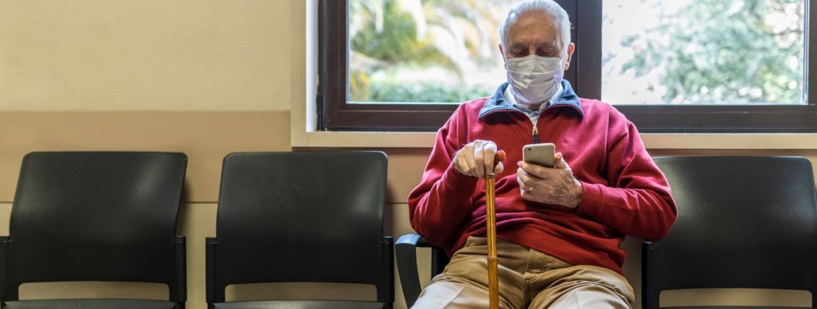 Elderly man wearing a face mask and carrying a stick sits in a hospital waiting room.