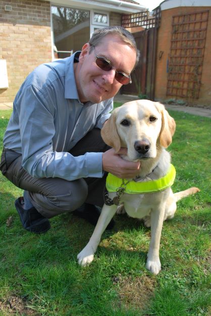 A photo of Paul in a garden, crouched down next to his guide dog Wanda, who is sat on the grass. Paul is wearing a blue shirt and grey trousers. Wanda is a blonde Labrador who is wearing her florescent harness.
