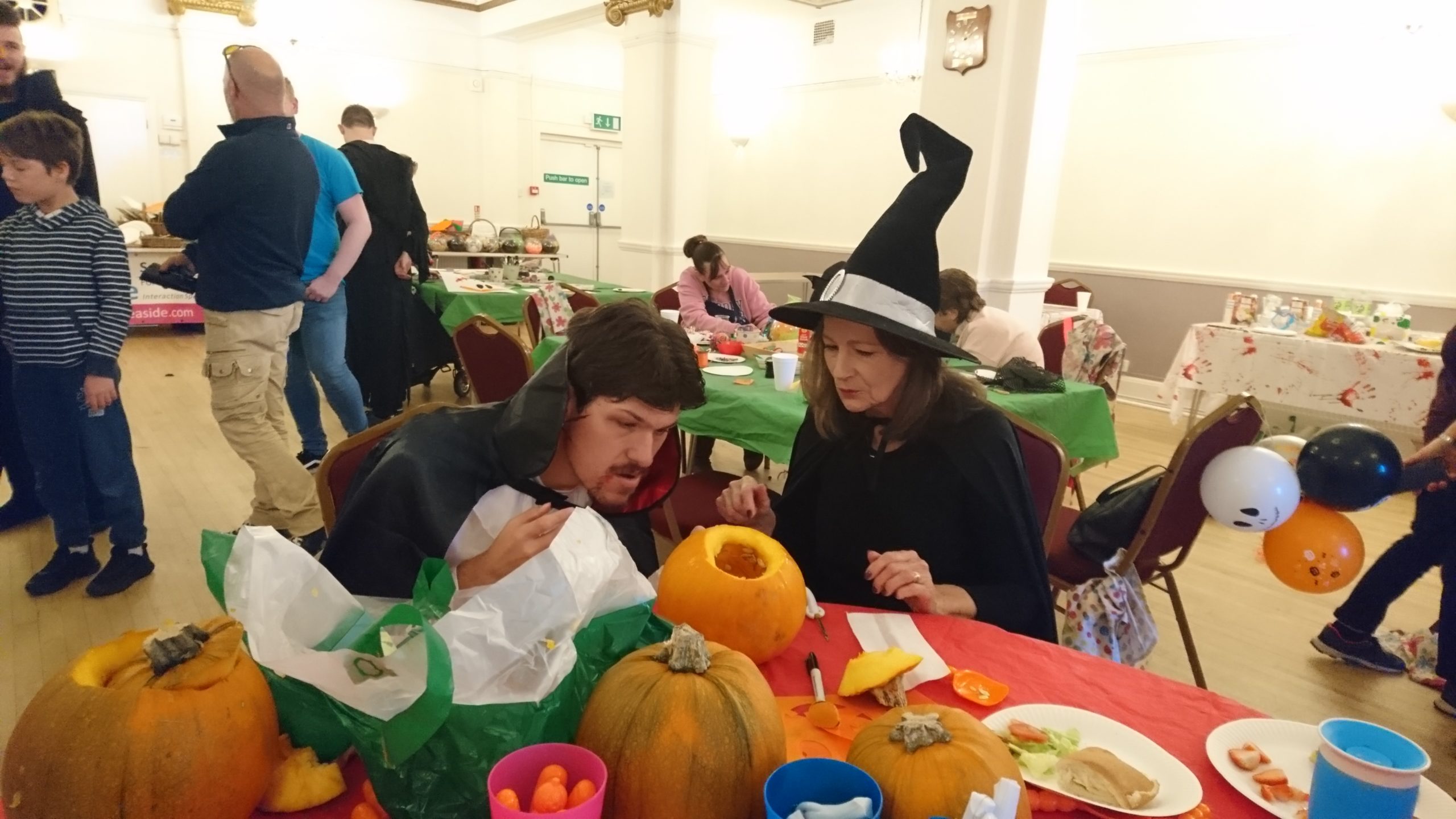 A woman wearing a witch hat for Halloween and a man hand carving pumpkins