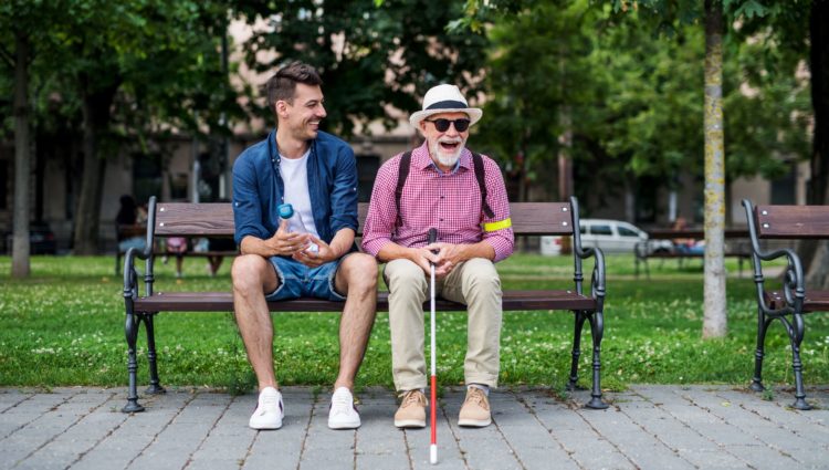 A young man and an older blind one siting on a bench and laughing